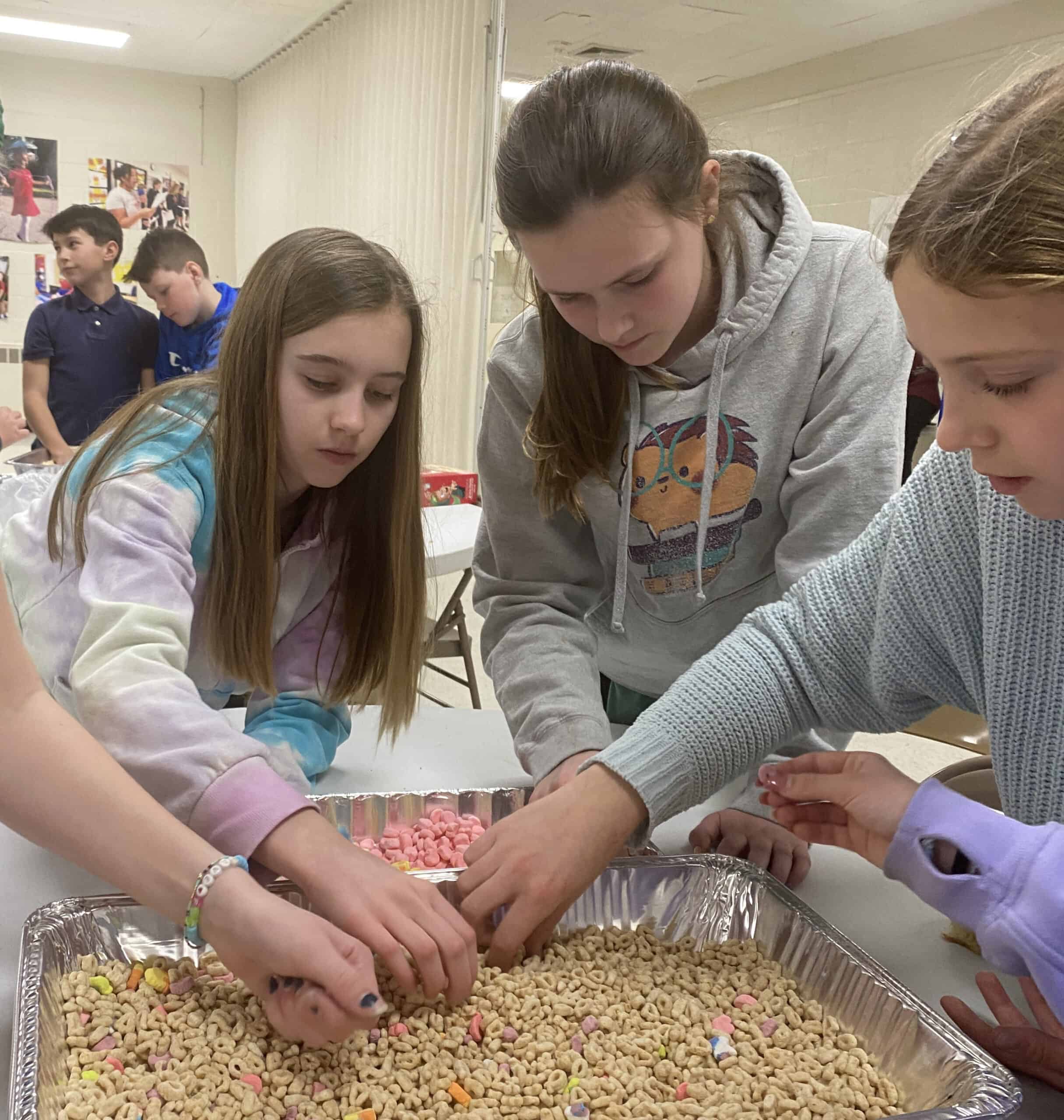 Three children sort colorful cereal pieces in large trays, while two other kids stand in the background near a decorated wall.