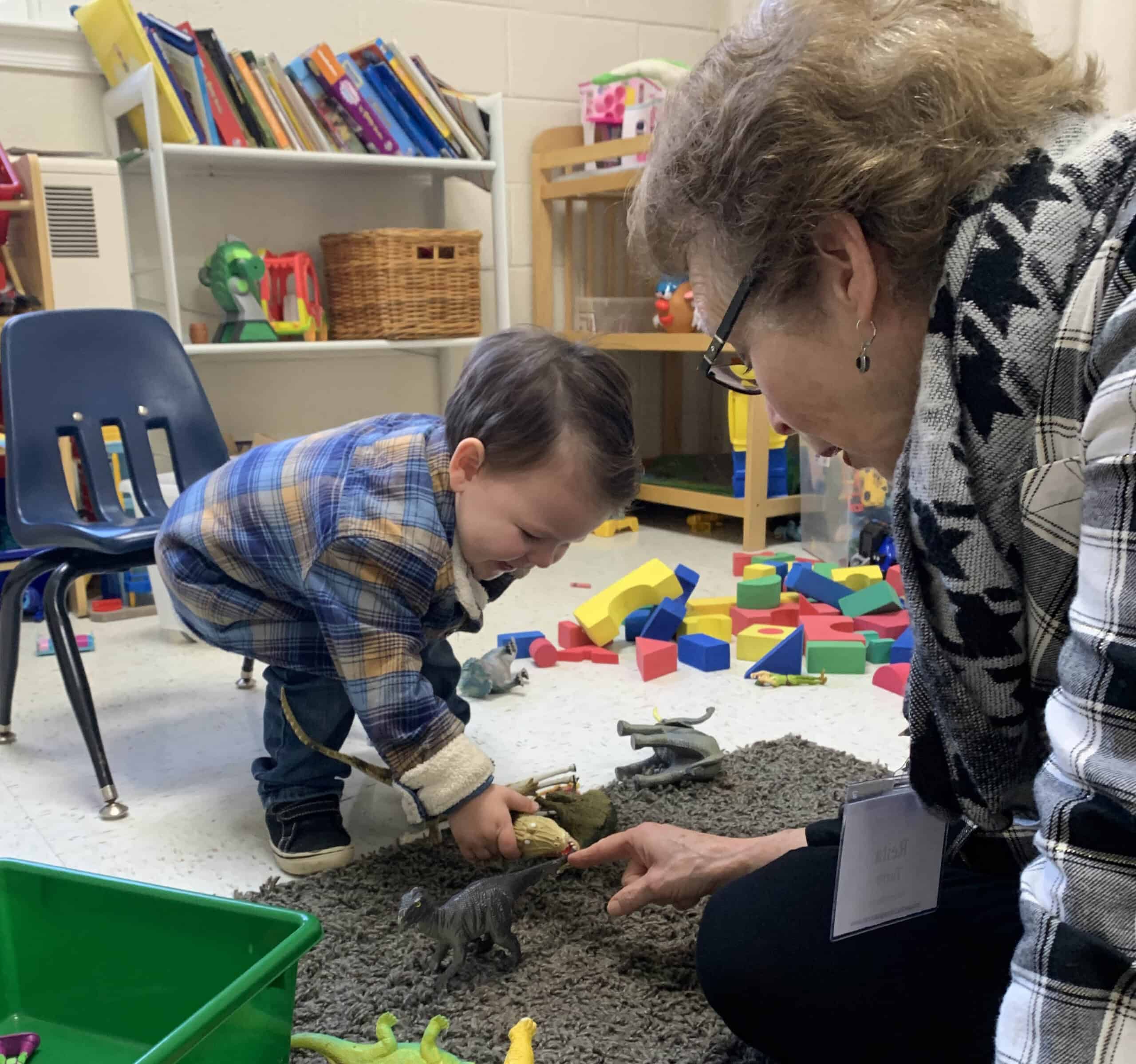 A child and person play with toy dinosaurs on a carpet in a colorful, book-filled classroom setting.