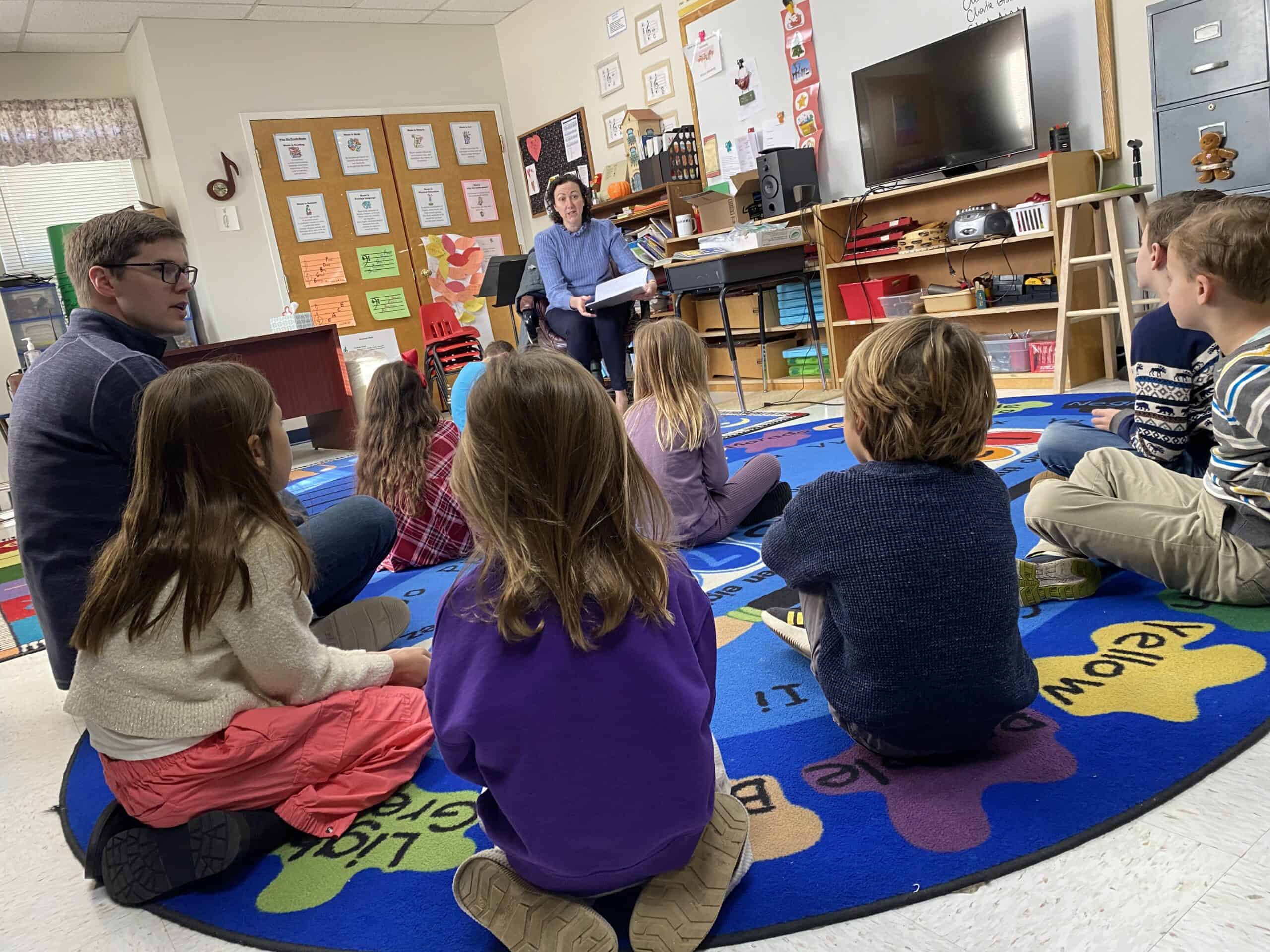 Children sitting on a colorful rug in a classroom, listening to a teacher reading a book.